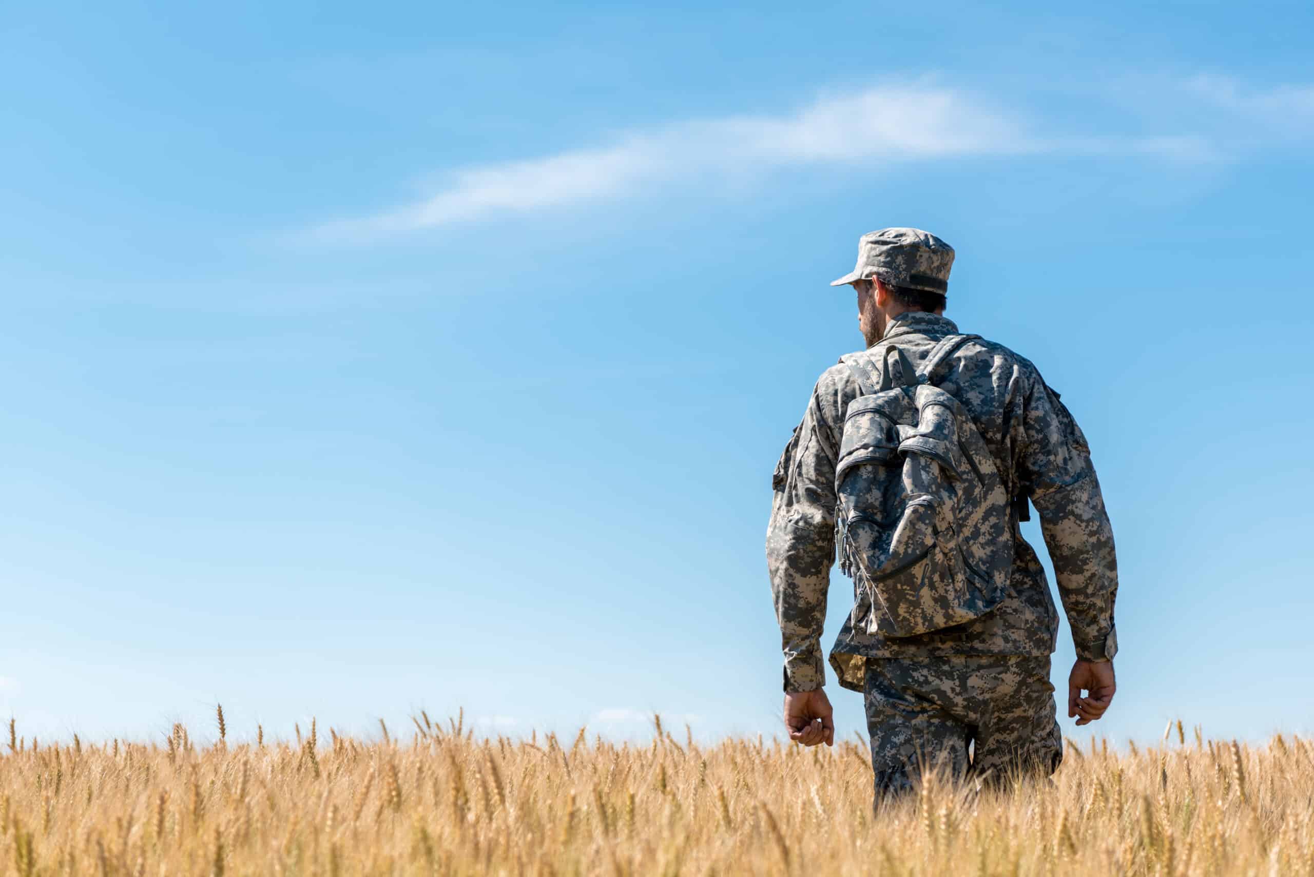 soldier in military uniform with backpack standing in field with golden wheat